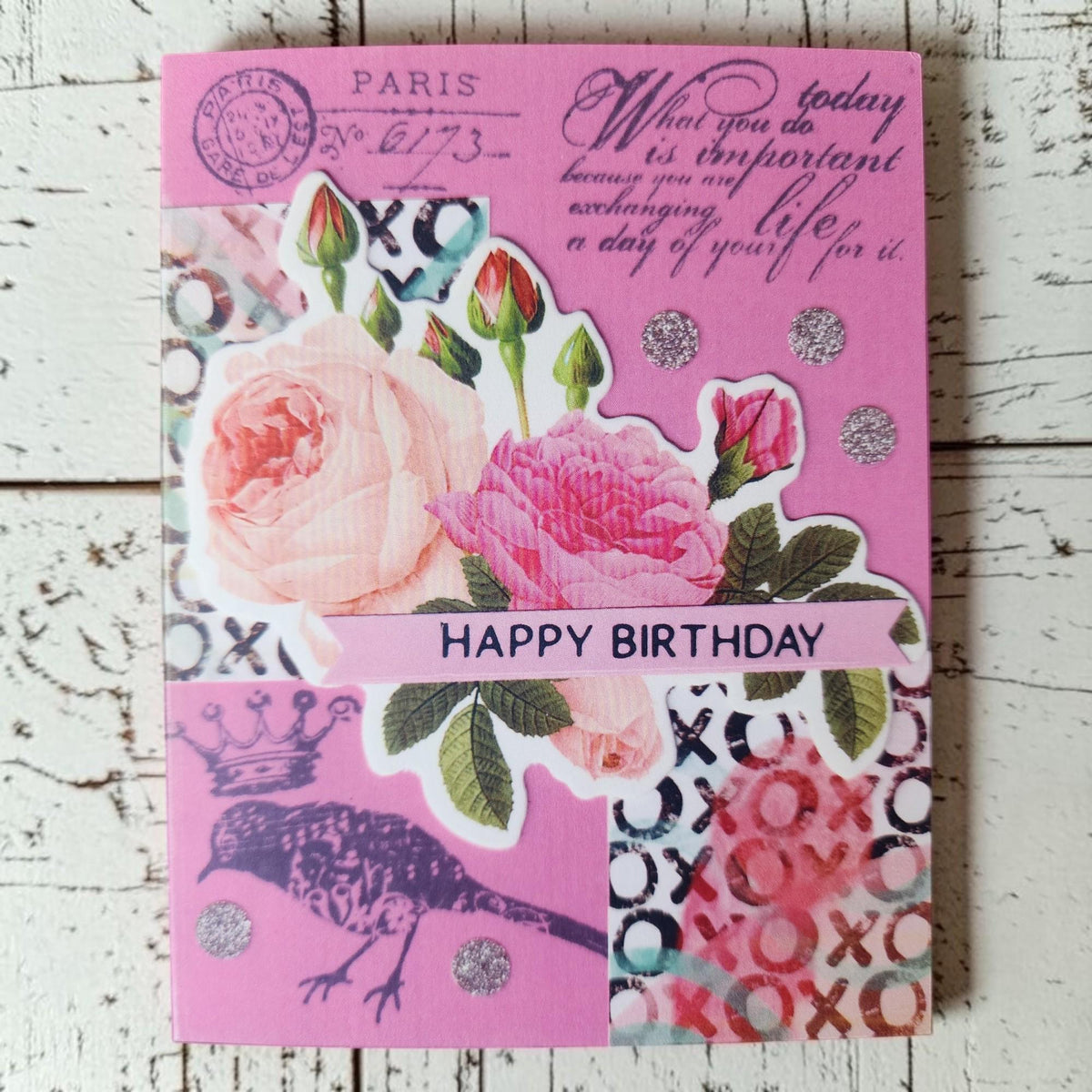 A pink handmade birthday card with floral and vintage-style graphics, including roses, postmark elements, script text, a bird silhouette, and 'HAPPY BIRTHDAY' printed across the center, photographed on a whitewashed wood surface.