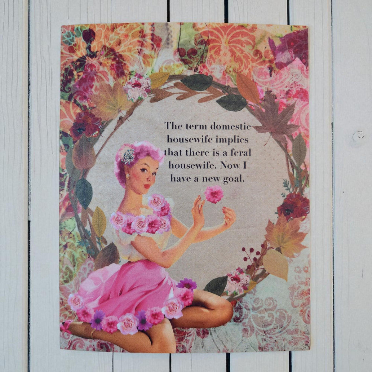 A vintage-style illustrated print or card featuring a woman in a pink dress seated among floral wreath elements, with black text in the center reading, 'The term domestic housewife implies that there is a feral housewife. Now I have a new goal.